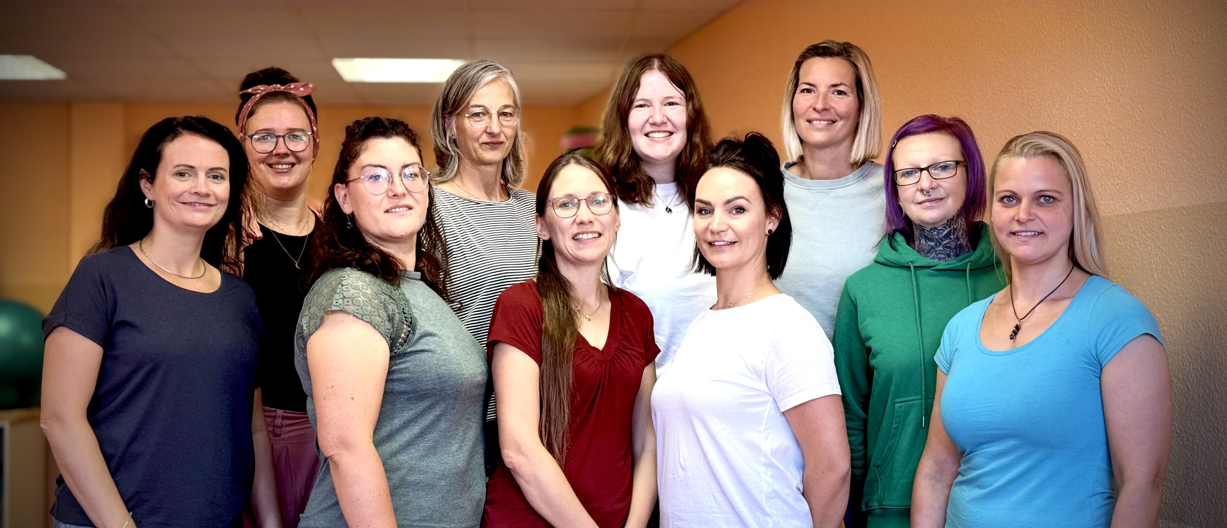 Zehn Frauen verschiedenen Alters lächeln freundlich in einem hellen Raum mit orangen Wänden