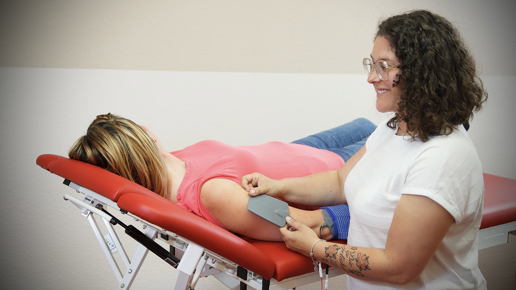 Eine lächelnde Frau mit Locken behandelt eine Patientin auf einer roten Liege