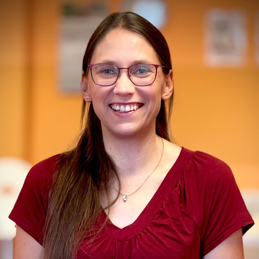 Eine lächelnde Frau mit Brille und langen braunen Haaren trägt ein burgunderrotes Oberteil