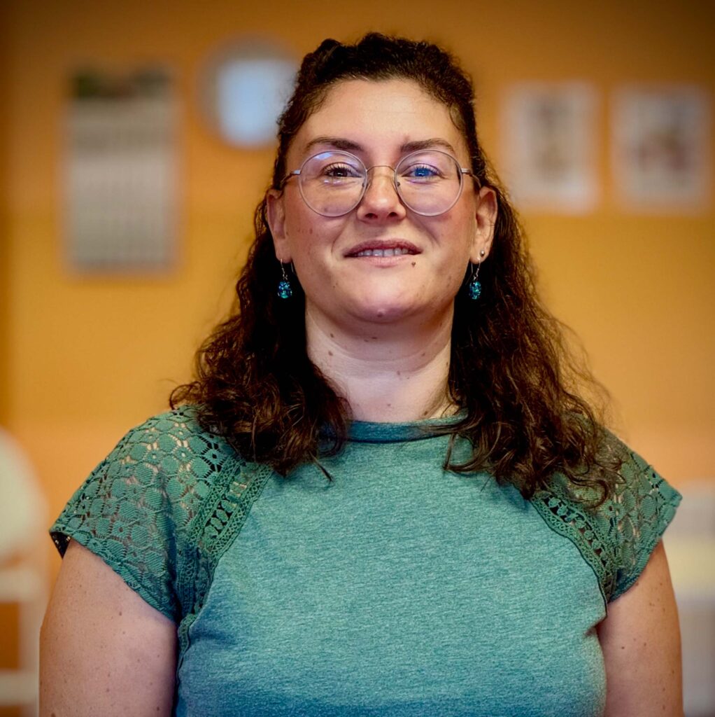 Eine Frau mit lockigem braunen Haar und Brille lächelt freundlich in grünem Oberteil