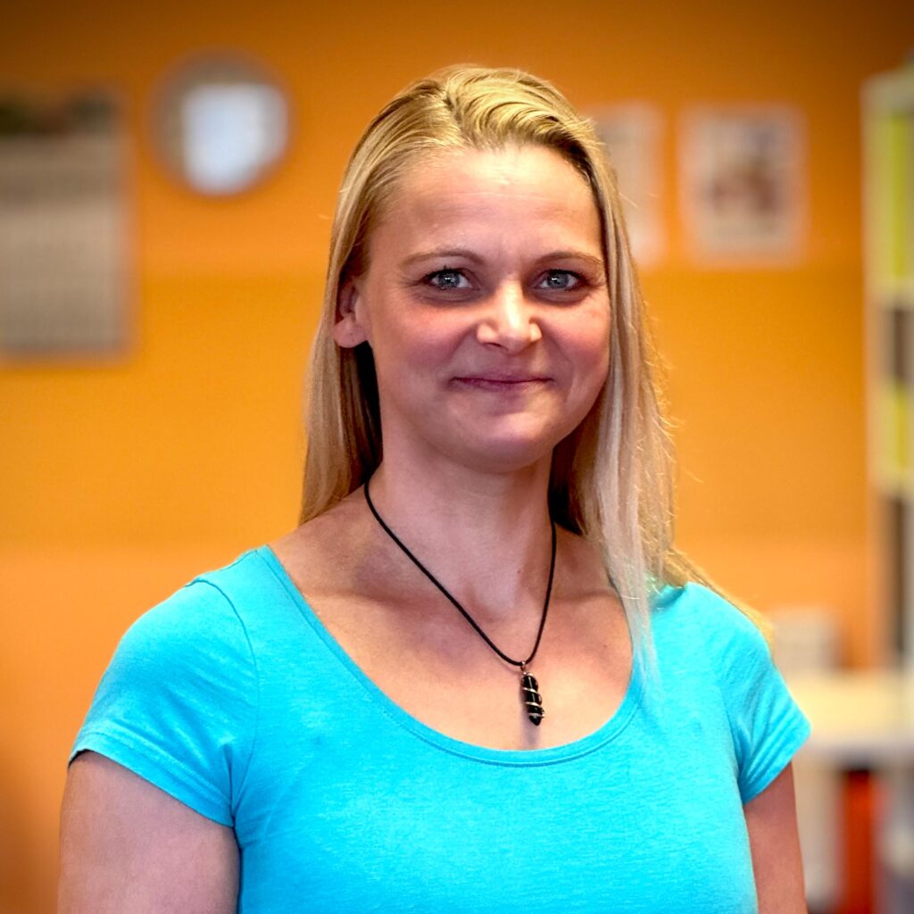 Eine lächelnde blonde Frau im blauen Shirt mit Kette steht vor orangefarbenem Hintergrund