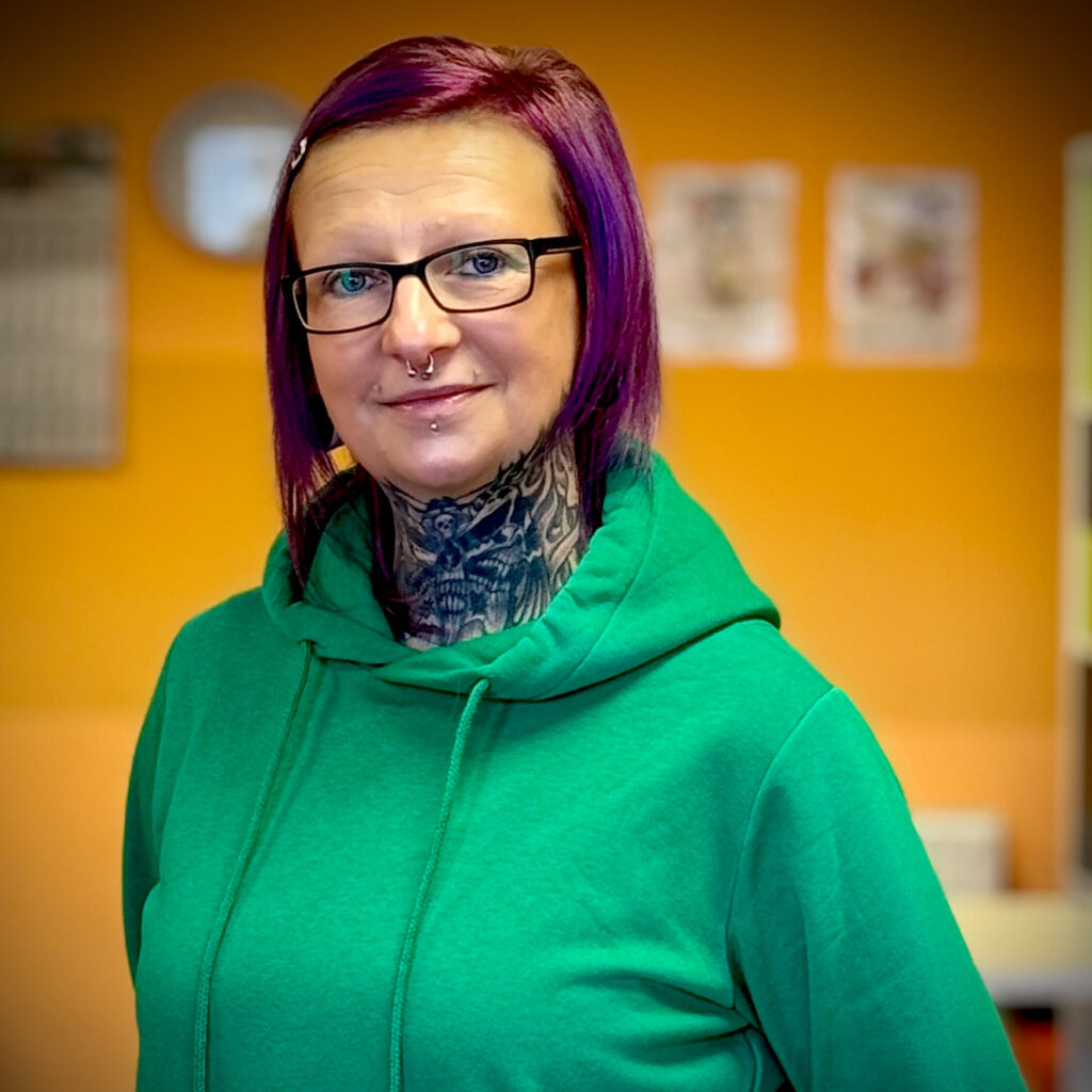 Frau mit lila Haaren und Brille lacht freundlich im grünen Hoodie vor oranger Wand