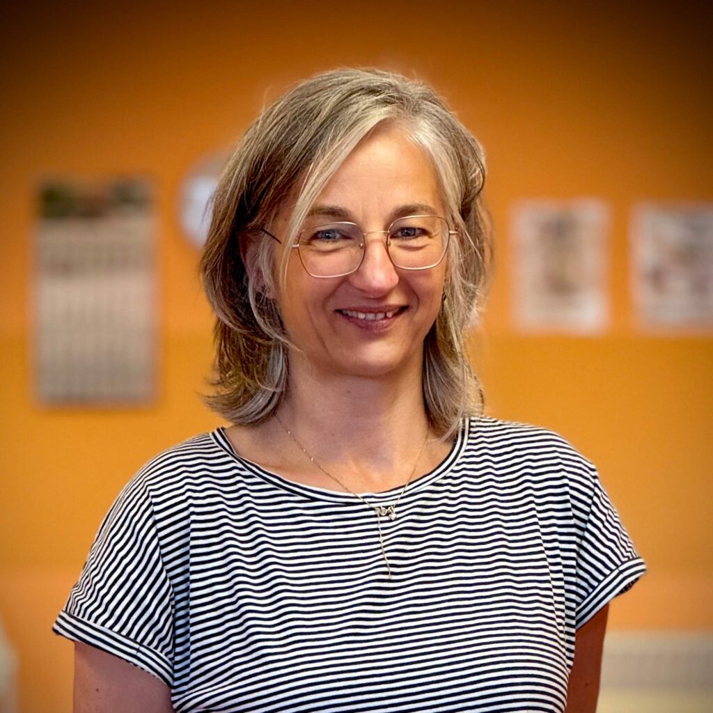 Eine freundlich lächelnde Frau mit Brille und graublondem Haar vor orangefarbenem Hintergrund