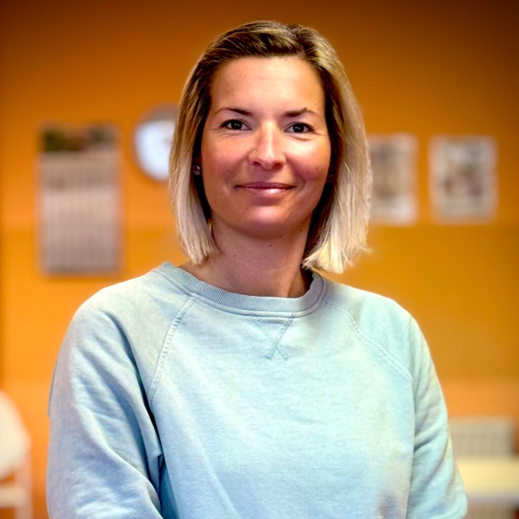 Eine Frau mit kurzem blondem Haar lächelt freundlich vor einer orangenen Wand