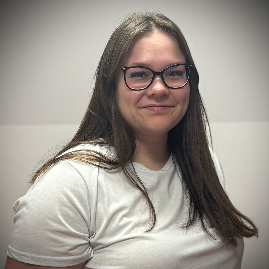 Eine junge Frau mit langen braunen Haaren lächelt freundlich und trägt eine Brille sowie ein weißes T Shirt