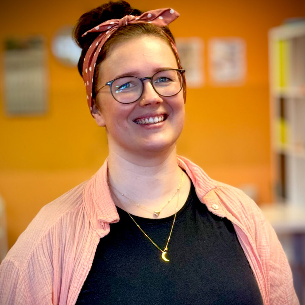 Eine junge Frau mit Brille lächelt freundlich und trägt ein rosa Stirnband und Hemd