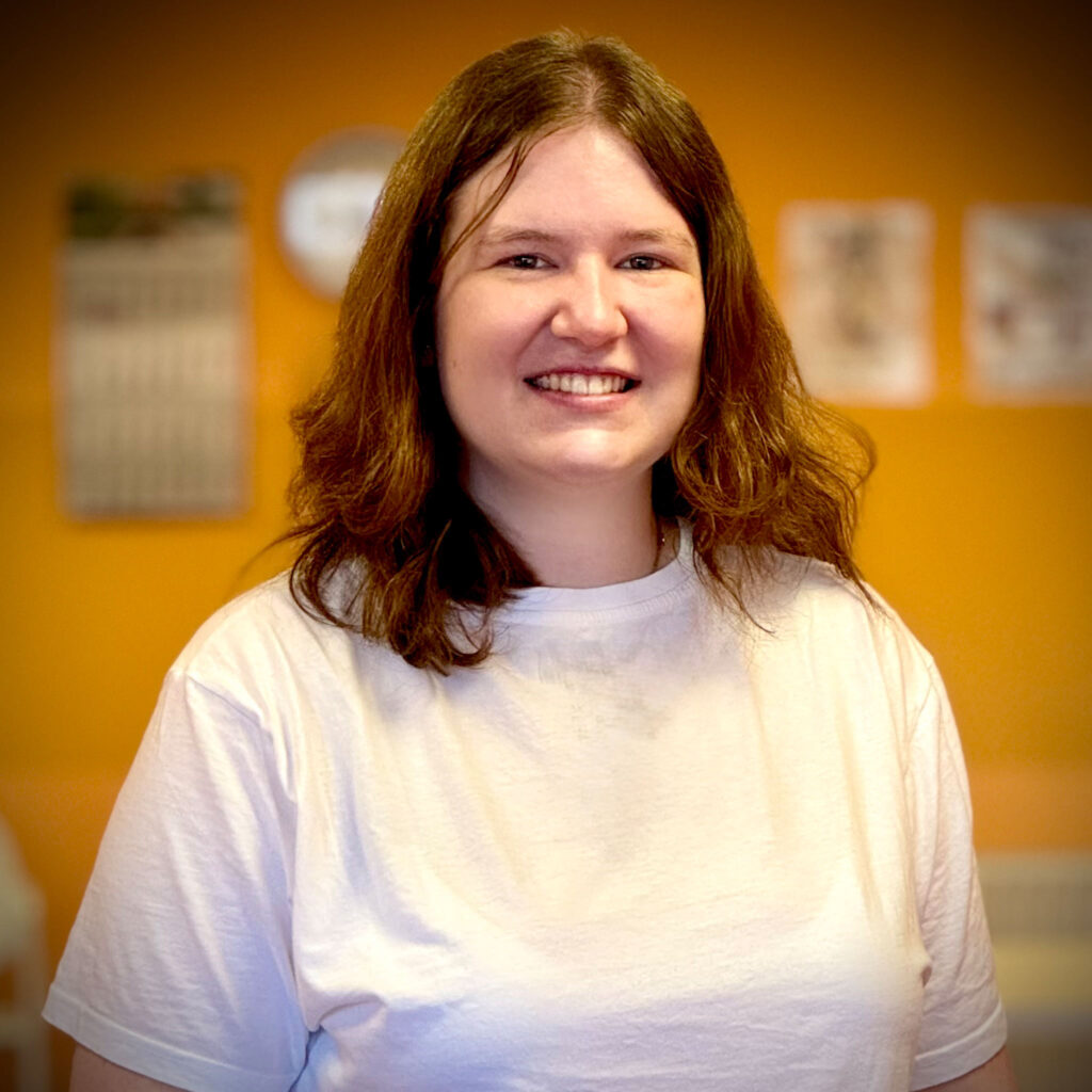 Junge Frau mit langen braunen Haaren lächelt freundlich im weißen TShirt vor orangefarbener Wand